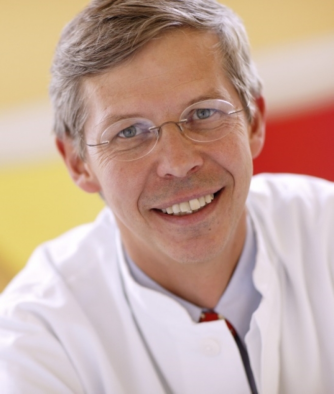 The photo shows a middle-aged man with a fair complexion, graying, short-cropped hair and light blue eyes. He wears thin, metallic glasses and a white apron, suggesting that he may be a doctor or scientist. He has a friendly, broad smile and looks happy. The background shows fuzzy colored areas in shades of yellow and red.