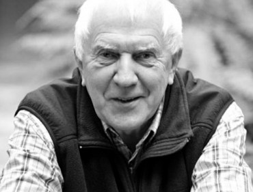 An elderly man in a vest and plaid shirt sits on a park bench, surrounded by greenery.