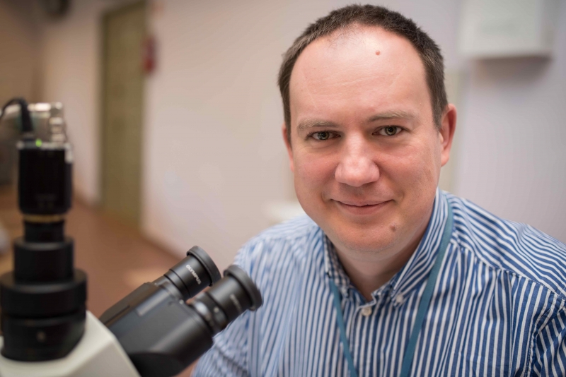 The photo shows a man with a fair complexion, short dark hair and a gentle smile. He wears a blue and white striped shirt and has a badge hanging around his neck. He is sitting at a microscope, suggesting that he is working in a laboratory or engaged in scientific research. In the background you can see the fuzzy interior of a room, probably a research or medical facility.