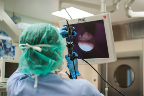 A surgeon, seen from behind, is performing an endoscopic procedure, holding the endoscope with gloved hands. The surgeon is wearing a green surgical cap and a blue surgical gown. The endoscope’s view is displayed on a monitor in front of the surgeon, showing the inside of the patient's body. The setting is a sterile operating room with medical equipment visible in the background
