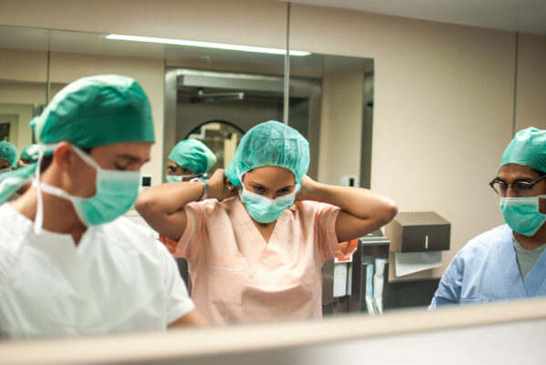 Three healthcare professionals are seen in a hospital setting, wearing surgical scrubs, masks, and caps. The person in the center, dressed in pink scrubs, is adjusting their surgical cap, while the others are focused on their tasks. They appear to be preparing for a medical procedure, with a reflection visible in the mirror behind them.