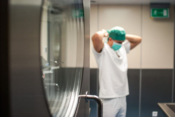 A medical professional in a surgical environment, seen through a reflective surface, adjusting their surgical cap. The setting is a sterile, modern medical facility, with the professional preparing for a procedure.
