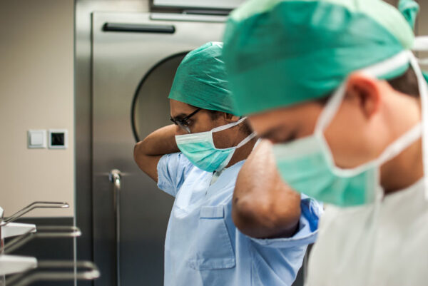 Two healthcare professionals wearing surgical scrubs and masks, focusing on adjusting their caps. The environment is a well-equipped medical facility, likely a hospital, with stainless steel equipment and clean surfaces.