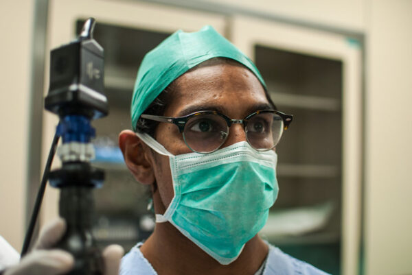 A close-up of a healthcare worker in surgical attire, including a green cap and mask, focusing intently on a task. The image captures the professional's concentration and the sterile environment of the operating room.