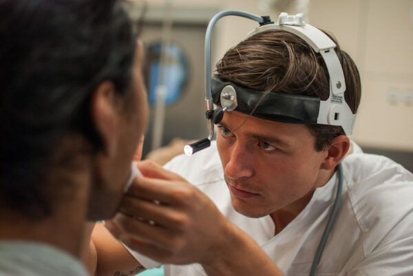 A focused medical professional wearing a headlamp and surgical attire, examining a patient closely. The scene reflects the precision and care involved in medical procedures, with the professional displaying intense concentration.