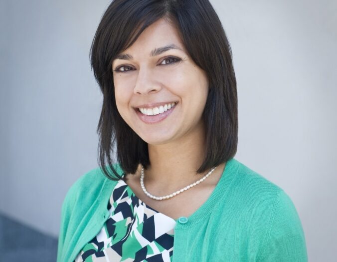 A smiling professional woman wearing a green cardigan and a patterned dress. She has dark hair styled into a neat bob and stands against a plain background, exuding warmth and approachability.