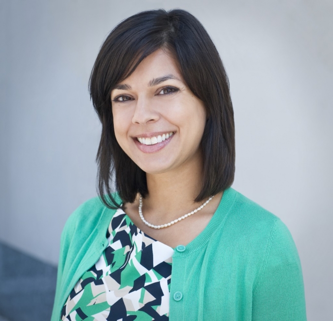 A smiling professional woman wearing a green cardigan and a patterned dress. She has dark hair styled into a neat bob and stands against a plain background, exuding warmth and approachability.