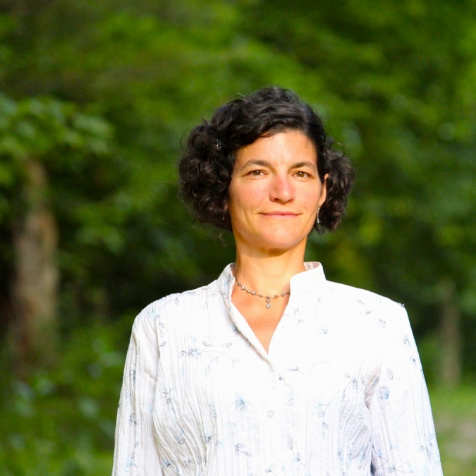 A woman with short hair stands confidently in front of a backdrop of lush green trees, embodying a serene outdoor moment.