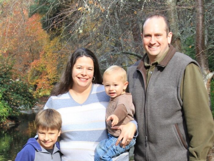 A family stands together smiling in front of a serene pond, capturing a joyful moment in nature.