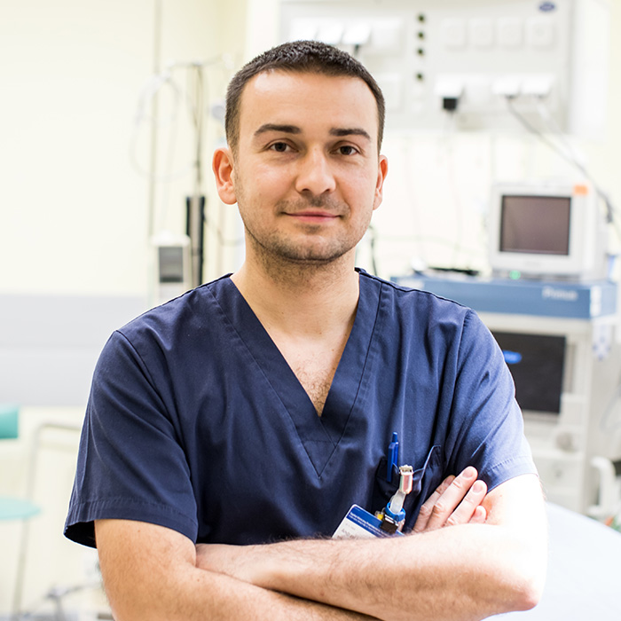A young medical professional in navy blue scrubs, standing confidently with arms crossed, with medical equipment visible in the background.