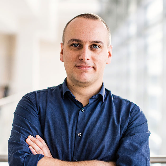 A headshot of a man with crossed arms. He is dressed in a dark blue shirt and stands in a modern, bright setting with a slight smile.