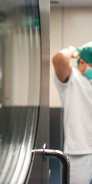 A healthcare worker in surgical scrubs and a green cap adjusts a mask, preparing for a procedure. The scene is set in a sterile medical environment with stainless steel equipment and clear lighting.