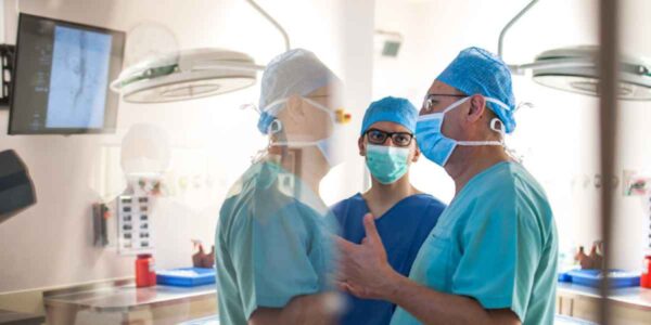 Three medical professionals in a hospital operating room, dressed in surgical scrubs, masks, and caps. They are engaged in a discussion, with a medical monitor visible in the background. The scene reflects a collaborative environment typical of a surgical team preparing for or discussing a procedure.