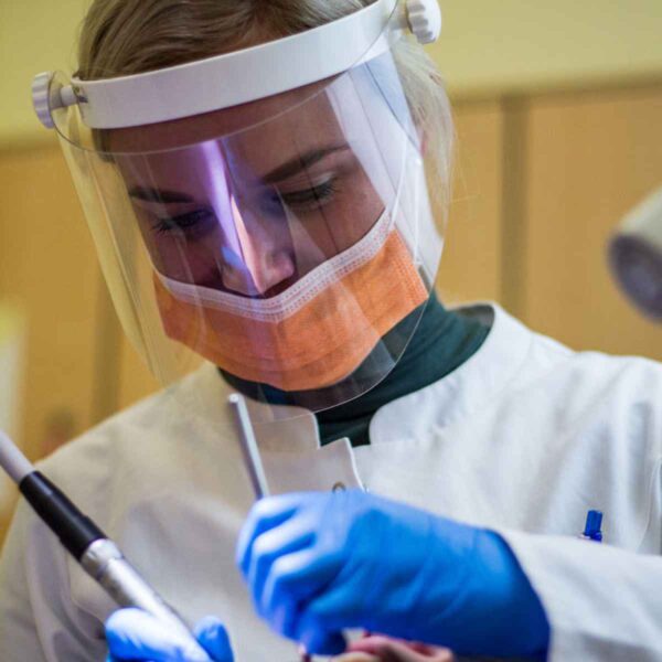 A close-up of a female medical professional wearing a face shield, mask, and gloves, focused on performing a dental or medical procedure.