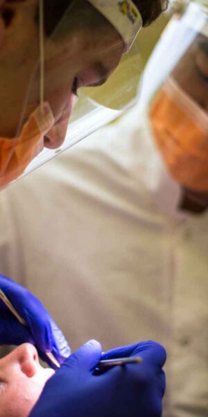 A close-up view of a medical professional with a face shield and orange mask, concentrating on a dental procedure. Another professional stands behind, observing.