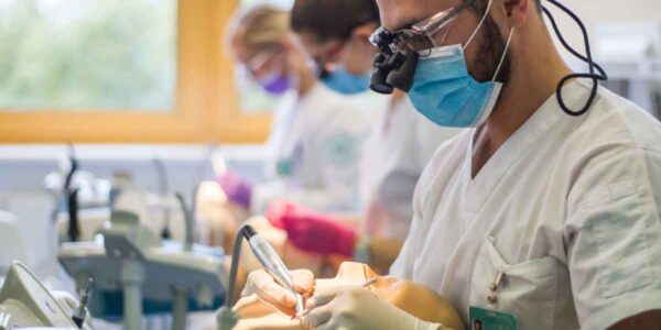 A dentist wearing magnifying glasses and a mask, using a dental drill on a mannequin. Other professionals in the background are blurred, also engaged in procedures.