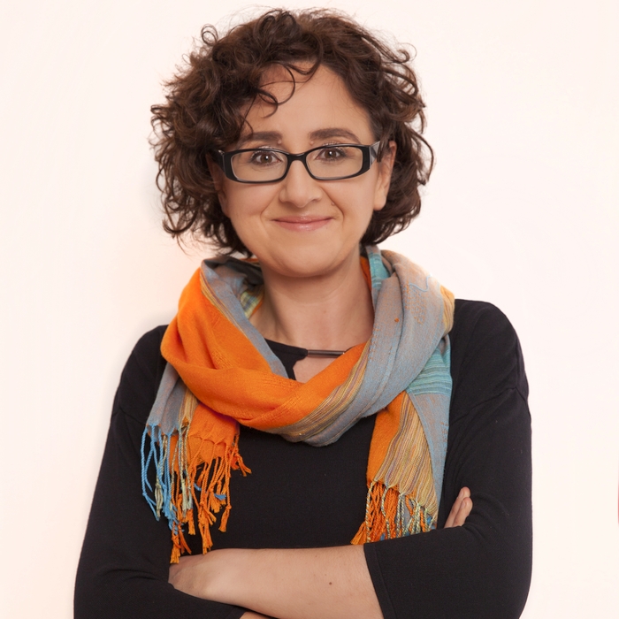 A woman with curly hair, glasses, and a friendly smile, wearing a colorful scarf over a black top, standing confidently with her arms crossed.
