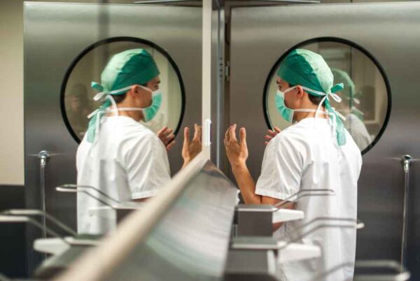 A surgeon, wearing a green cap and mask, is preparing for surgery by thoroughly washing their hands before entering the operating room. The background shows a sterile medical environment with metal doors leading to the operating room. The surgeon's reflection is visible on the glass surface to the left.