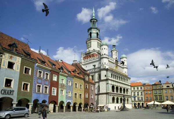 A vibrant town square featuring colorful buildings and a large, historic clock tower with multiple domes. The square is lively with outdoor seating and people walking, capturing the charm and cultural richness of the area.