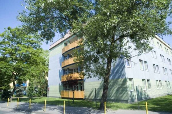 A residential building with a modern design, featuring balconies and surrounded by trees. The building is painted in soft pastel colors, creating a calm and inviting atmosphere in what appears to be a peaceful neighborhood.