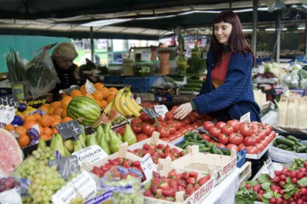 A bustling farmers market with stalls filled with fresh fruits and vegetables. A woman is selecting produce, surrounded by a colorful array of strawberries, tomatoes, cucumbers, and other fresh items, capturing the lively and community-oriented atmosphere of the market.
