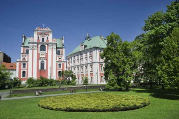 A church with a historic facade, viewed from a parking lot. The church is surrounded by green trees and the sky is clear and blue.