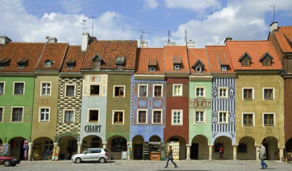 A street view leading up to a large Baroque church facade painted in red and white, with a cobblestone street and surrounding buildings in pastel colors.