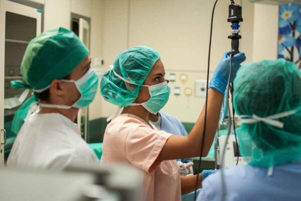 A group of healthcare workers in a hospital setting, wearing scrubs, masks, and surgical caps. One of them is adjusting an IV drip, while others observe or assist. The room is equipped with various medical instruments and displays, indicating an active medical environment.