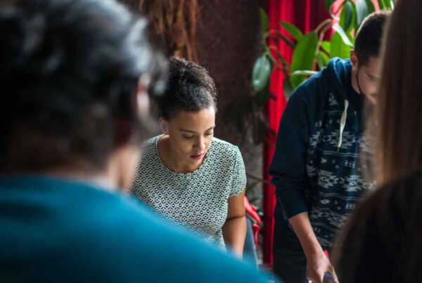 A candid photograph of a group of people working together. A woman in the center is focused on her task, wearing a patterned shirt, with other individuals blurred in the foreground. The setting is informal and likely a creative or collaborative workspace.