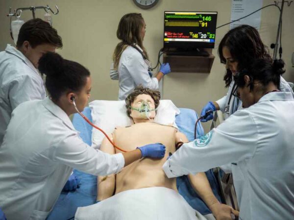 A medical simulation scene with a group of healthcare students practicing on a mannequin. The students, dressed in white lab coats and gloves, are engaged in a realistic training exercise, checking vitals and administering care while monitoring the patient’s condition on a screen.