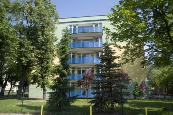 A pastel green and yellow residential building with balconies, surrounded by lush green trees and a fence in the foreground.