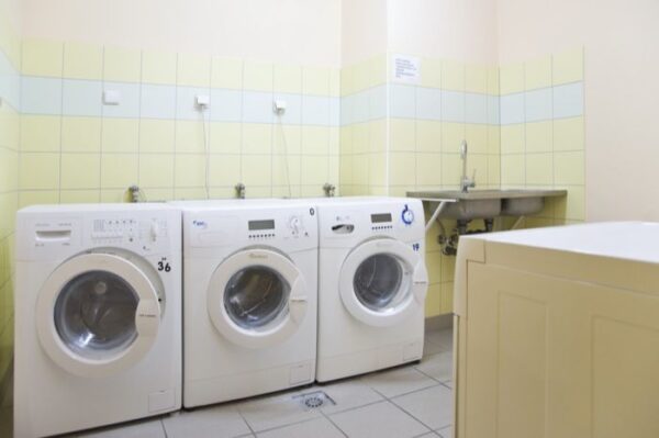 A clean laundry room equipped with three front-loading washing machines, tiled floors, and a stainless steel sink area with taps.