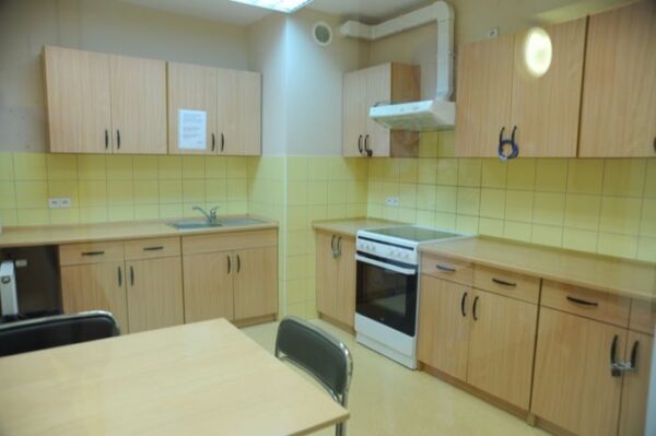 A communal kitchen with wooden cabinets, a white stove, and tiled walls in a pale yellow and green color scheme. There is a table with chairs in the center of the kitchen.