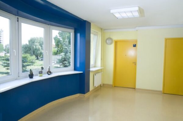 A common hallway painted in yellow and blue tones, with large windows allowing natural light to stream in. The doors are painted in bright yellow.