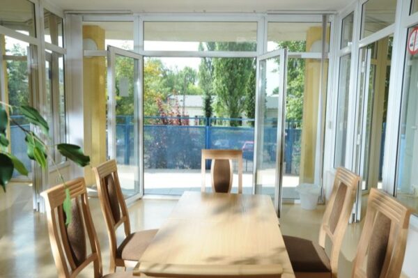 A lounge area with large windows and sliding glass doors leading outside. The room features a wooden dining table with chairs and has plenty of natural light.