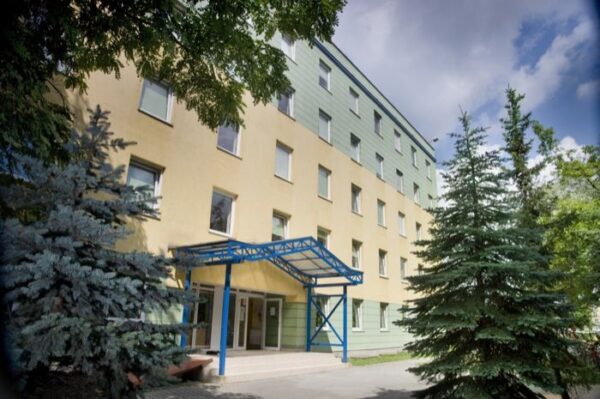 The exterior of a residential building with green and yellow walls and a blue metal entrance structure. The building is surrounded by trees.