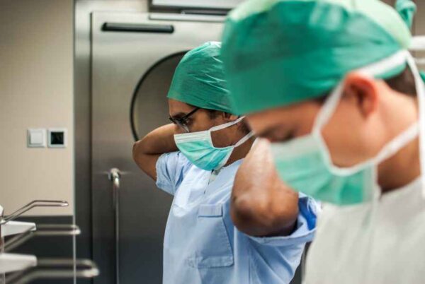 Two healthcare professionals wearing surgical scrubs and masks, focusing on adjusting their caps. The environment is a well-equipped medical facility, likely a hospital, with stainless steel equipment and clean surfaces.