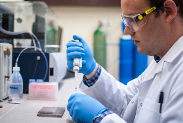 A scientist or laboratory technician working in a lab, using a pipette while wearing protective gear, including gloves and safety glasses. The setting is a research or clinical laboratory, with scientific instruments visible in the background.