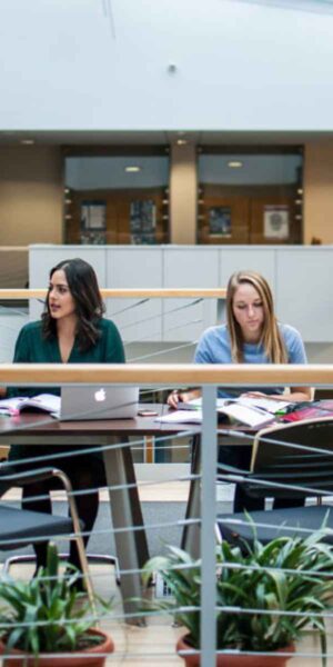 Two students collaborate on laptops in a bright atrium, surrounded by natural light and modern architecture.
