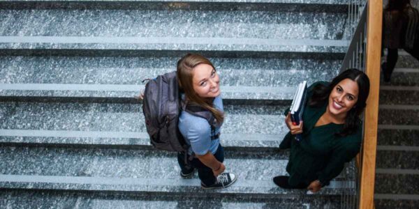 Two students stand on a staircase, books clutched in their hands, symbolizing academic collaboration and friendship.