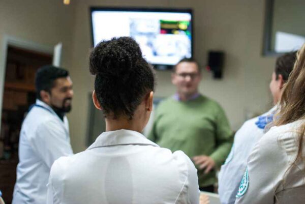 A group of students, some in white lab coats, are gathered around a person giving a presentation. The focus is on the back of a student with curly hair, while a blurred screen is visible in the background.