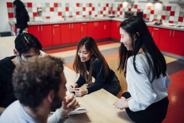 A group of students in a brightly lit kitchen area, laughing and conversing around a table. The red cabinets and checkered backsplash create a casual and lively setting.