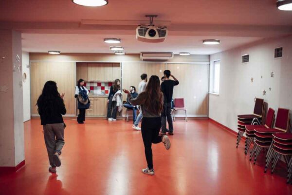 A group of students casually engaging in a large open room with orange flooring and light-colored walls. The mood is informal, as some are walking or playfully moving while others chat.