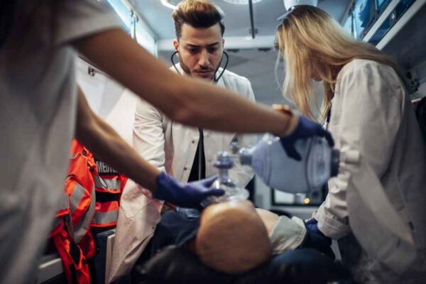 A medical simulation in progress inside an ambulance. A male student closely monitors the dummy while another student applies a manual resuscitator to simulate providing ventilation.