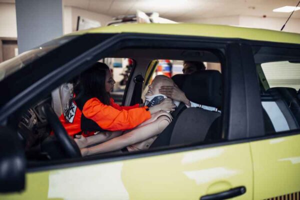 A simulated rescue operation where a paramedic in an orange jacket provides care to a dummy in a car. The scene captures the practical application of emergency procedures.