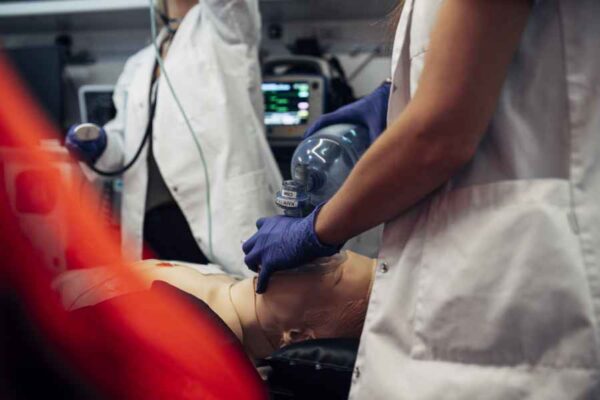 A close-up of a simulation exercise where a medical student performs manual ventilation using a bag-valve mask on a dummy. Monitors in the background display vital signs, adding realism to the practice.