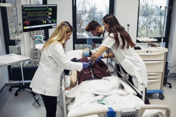 Medical students in white lab coats working on a medical dummy in a hospital room. They are conducting a simulated emergency, with a monitor displaying vital signs in the background. The students are performing chest compressions and using medical equipment to stabilize the dummy.