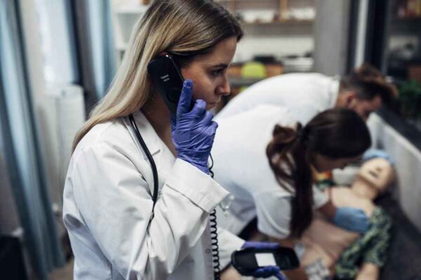 A medical student in a white coat talking on a phone while others perform chest compressions on a medical dummy. The scene appears intense as students manage a complex emergency scenario in a simulated environment.