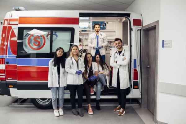 A group of medical students posing in front of an ambulance. They are dressed in white lab coats, with a mix of smiles and serious expressions, symbolizing teamwork and readiness for medical emergencies.