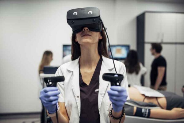 A student in a lab coat using a virtual reality headset while holding controllers. The setup is likely part of a medical training module, with others working on simulations in the background.